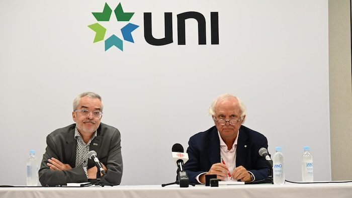 Pierre-Marcel Desjardins and Camille Theriault sit next to each other at a table during a press conference.  Above their heads is the UNI logo. 