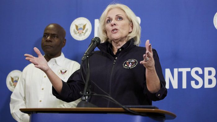 A blonde woman, standing in front of a desk, arms outstretched, wearing a shirt bearing the badge of the American Transportation Security Administration.  A black man is behind her, to her right. 